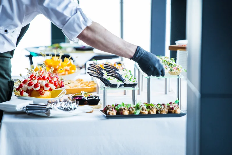 A hospitality staff member in a white chef's jacket and black gloves is arranging gourmet canapés and small bites on a buffet display table.